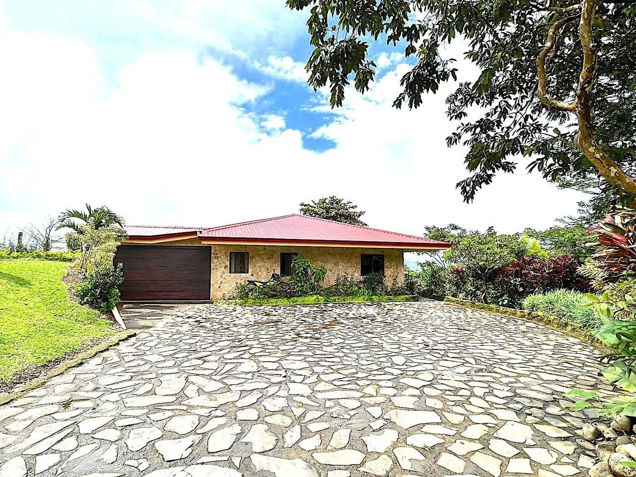 Single-story stone house with a red roof at the end of a flagstone driveway, framed by tropical gardens and trees