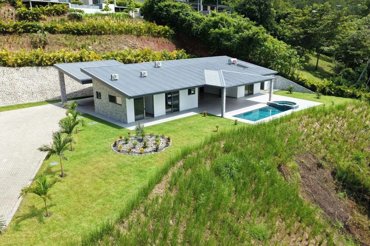 Aerial view of a new single-story home with a gray roof, private pool and jacuzzi on a landscaped hillside lot
