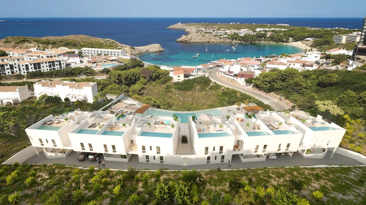 Aerial view of white apartment buildings with rooftop pools above a turquoise bay at Arenal d'en Castell, Menorca